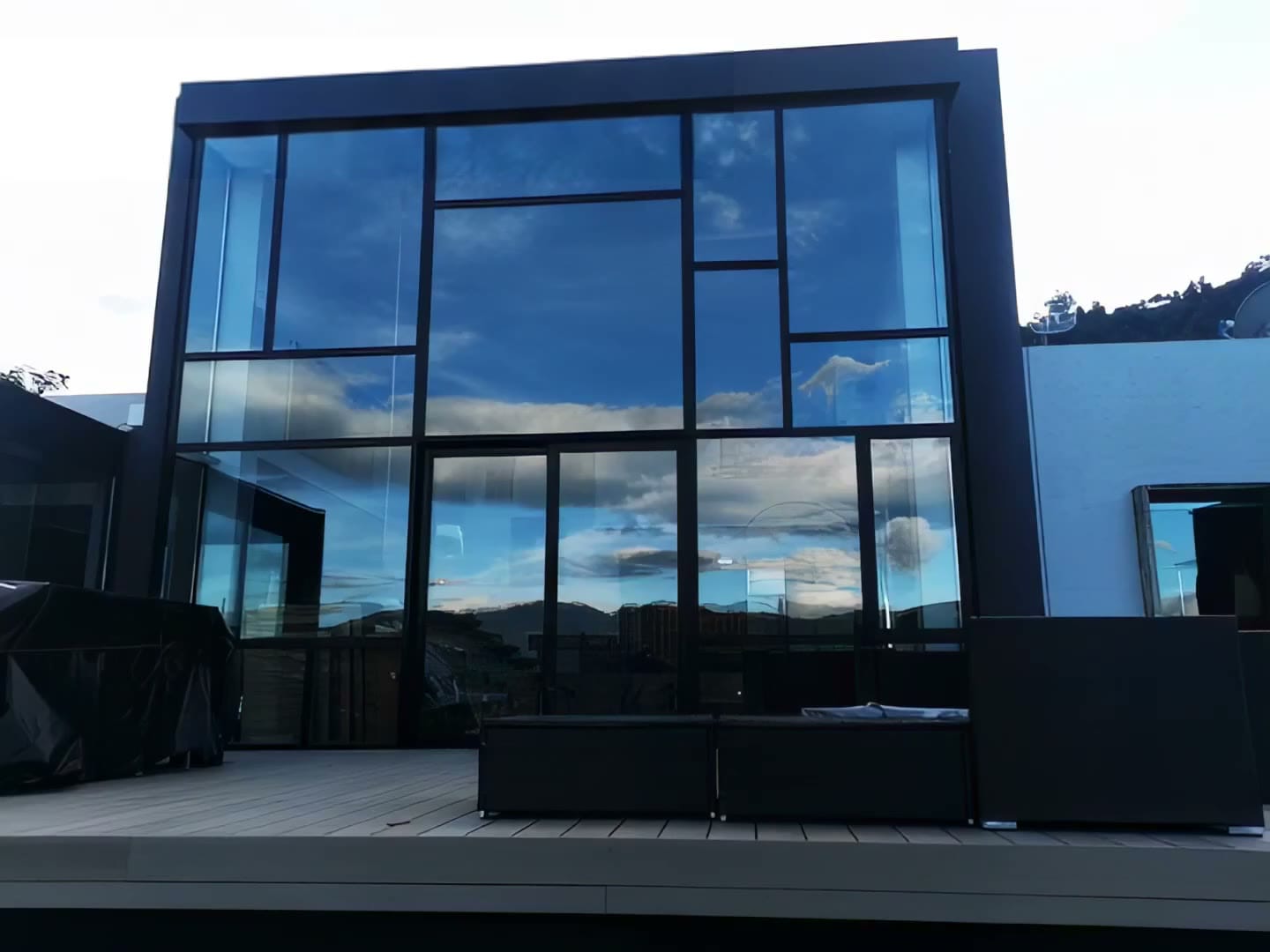 Fachada de vidrio con perfilería oscura que refleja el cielo y las montañas al atardecer.
